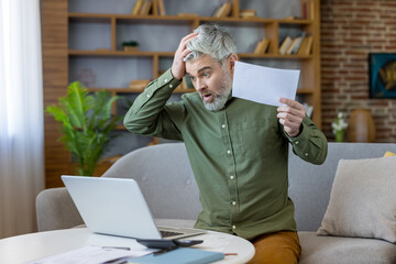 Mature man at home reacts in shock and stress reading online banking and bills on his laptop, facing debt and financial crisis while reviewing documents and paperwork in living room