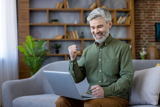 Mature man with grey beard celebrates winning news while working remotely on a laptop from his cozy home office sofa, smiling with excitement and relaxed confidence (160 chars)