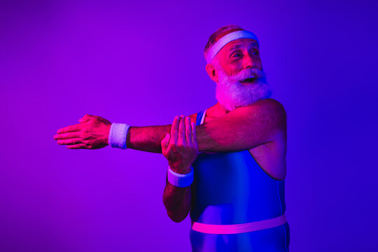 Male senior fitness model in retro blue and purple lighting stretching arms with bearded smile in vintage jumpsuit