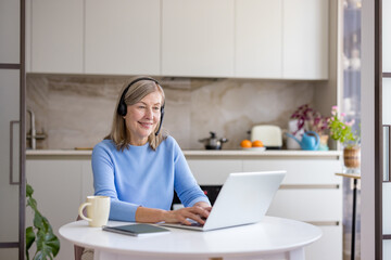 Smiling senior woman wearing a headset engaging in an online video call or remote work using a laptop computer from her modern kitchen, learning and connecting