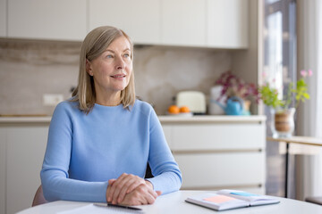 Senior woman is sitting at a white table in her modern kitchen, hands clasped, looking away thoughtfully while planning her future goals and daily tasks in an open notebook