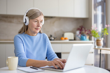 Senior woman concentrating on her laptop screen, wearing white headphones while typing on the keyboard and sitting at a round white table in a bright modern kitchen