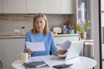 Senior woman at home kitchen table feeling stressed and confused while looking at multiple bills and financial documents, working on her laptop to manage personal finances