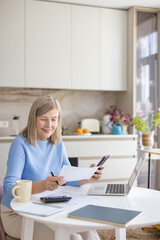Smiling senior woman managing finances, calculating expenses, and paying bills using a laptop,...