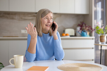 Senior woman in modern kitchen looks upset and gestures while on a frustrating phone call, conveying confusion, irritation and stressful miscommunication at home