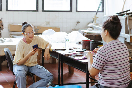 Art students using phone and chatting in studio, learning creative skills, collaborating on project, preparing artwork