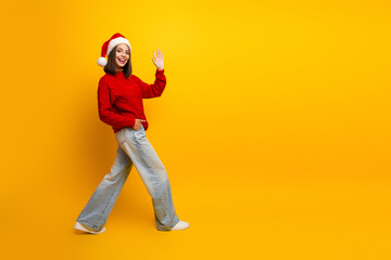 Playful Santa girl struts in a red sweater and jeans waving hello against a bright yellow backdrop for a cheerful holiday mood and everyday festive lifestyle