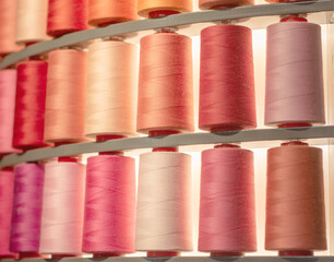Close-up of numerous large thread spools arranged in a warm color gradient of reds, pinks, and creams. Organized textile manufacturing or sewing supply display with backlighting.