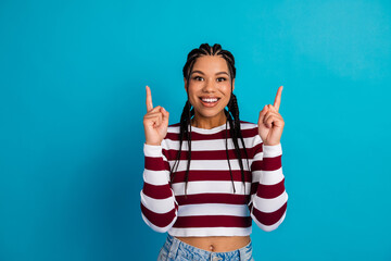 Young woman with braids in a striped top points upward against a blue background smiling brightly
