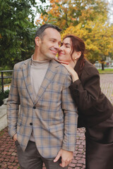 Happy smiling woman leaning affectionately on a stylish man's shoulder outdoors, surrounded by yellow and green autumn foliage
