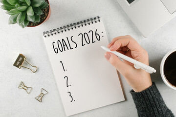 Woman's hand writing 2025 Goals in notebook. Laptop, home plant, cup of coffee on light table. Top view, flat lay
