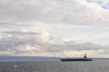 Aircraft Carrier On Calm Sea Under Cloudy Sky With Small Sailboat Nearby