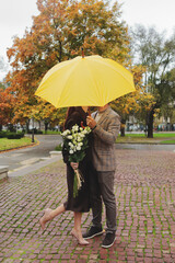 A romantic couple secretly kisses under a bright yellow umbrella while holding a bouquet of white flowers on a cobblestone street in autumn