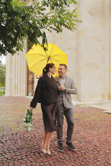 Happy elegant couple smiling at each other under a bright yellow umbrella, holding white roses on a red cobblestone street in autumn