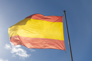 A vibrant shot of the Spanish national flag waving proudly in the bright sunlight, its yellow and red colors vivid against a deep blue sky scattered with soft white clouds.