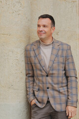 Happy, stylish middle-aged man in a plaid blazer and turtleneck, leaning casually against an old limestone wall and looking away