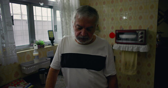 Elderly man standing in kitchen, looking down in contemplation, expressing solitude and quiet reflection in a domestic environment