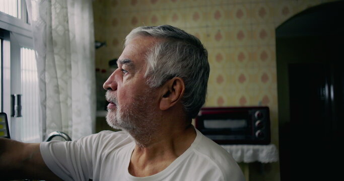 Elderly man gazing out of kitchen window with introspective expression, representing solitude, aging, and personal moments of reflection in everyday life