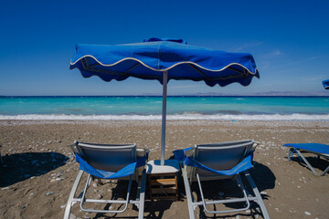 Row of empty sun loungers under blue beach umbrellas on a sunny pebble beach with calm turquoise sea in the background. Summer holiday concept with copy space for travel agencies.