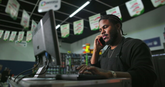 One Confident Black male store employee multitasking at counter, answering phone call while typing on computer, efficient retail work in busy shop environment