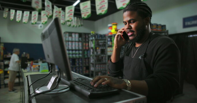 One Confident Black male store employee multitasking at counter, answering phone call while typing on computer, efficient retail work in busy shop environment