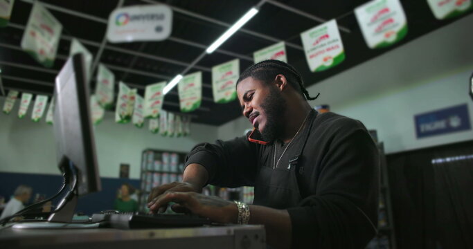 One Confident Black male store employee multitasking at counter, answering phone call while typing on computer, efficient retail work in busy shop environment - Powered by Adobe