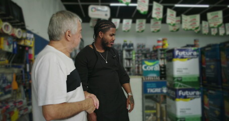 Obraz premium Older man and younger male worker pointing at paint cans in a hardware store aisle, discussing products, diverse teamwork in professional retail environment
