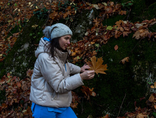 A woman kneels near a mossy rock, holding an autumn leaf as she explores the forest floor. The warm...