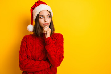 Woman in red sweater and Santa hat ponders holiday plans against bright yellow background for...