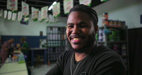 Confident male paint store assistant smiling with arms crossed in front of checkout counter, shelves of paint cans and supplies visible in background