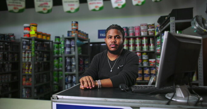 Man at paint store counter seated with hands clasped, wearing black apron, surrounded by colorful product shelves, exuding confidence and professionalism