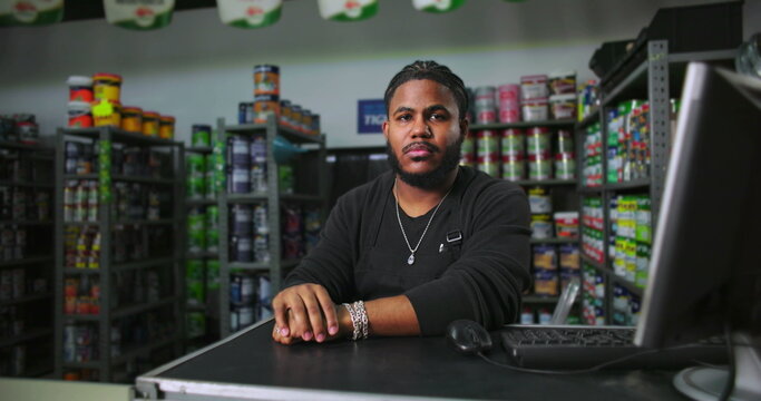 Man at paint store counter seated with hands clasped, wearing black apron, surrounded by colorful product shelves, exuding confidence and professionalism