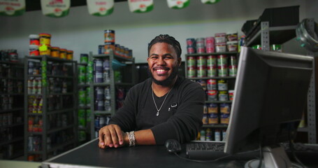 Smiling man working in paint store, sitting at counter with organized shelves of paint cans in background, professional and approachable worker at the desk