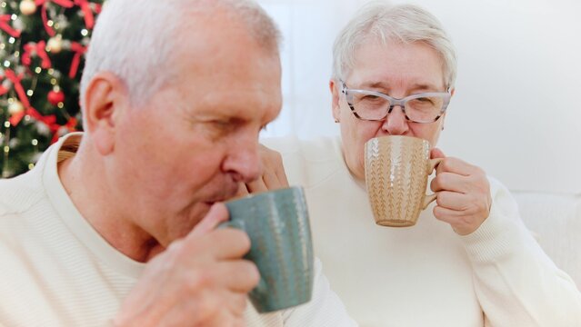 Elderly couple enjoying warm drinks tea together while celebrating New Year near a Christmas tree - Powered by Adobe
