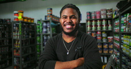 Man standing confidently at paint store counter with arms crossed, surrounded by paint cans and supplies, welcoming worker in professional retail setting