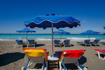 Row of empty sun loungers under blue beach umbrellas on a sunny pebble beach with calm turquoise sea in the background. Summer holiday concept with copy space for travel agencies.