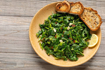 Cooked chicory greens sautéed in olive oil with garlic and hot pepper flakes - a traditional Italian side dish served with toasted bread and a slice of lemon. Rustic wooden background.