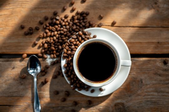 op-view flatlay of a cup of black coffee on a saucer with scattered roasted coffee beans and a small spoon on a rustic wooden table, warm natural light, no hands, no humans, cozy cafe atmosphere.