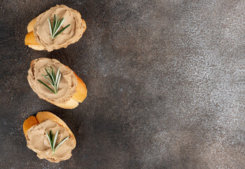 Three baguette slices with chicken liver pate and rosemary on dark rustic background. Top view. Copy space.