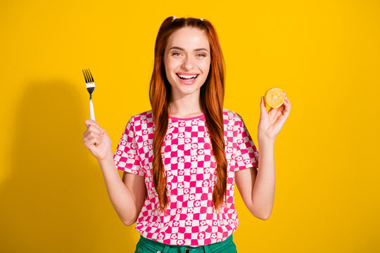 Young girl with pink checkered shirt holding fork and lemon against bright yellow background smiling happily