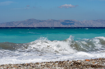 Turquoise sea waves crashing on pebble beach with mountains in background