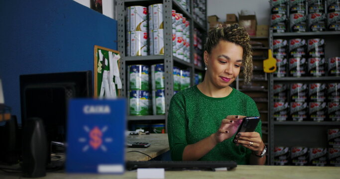 Young woman seated at a desk in a hardware store, smiling while checking her smartphone, professional retail business atmosphere with shelves of products