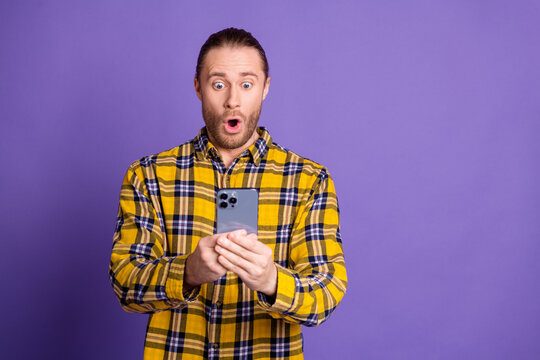 Young handsome man with smartphone surprised by screen wearing checkered yellow shirt against purple background - Powered by Adobe