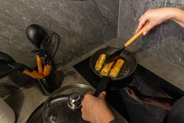 Person Frying Rolled Pancakes on Induction Stove. Hands flipping golden rolled pancakes in a frying pan on an induction cook top, with kitchen utensils visible in the background.