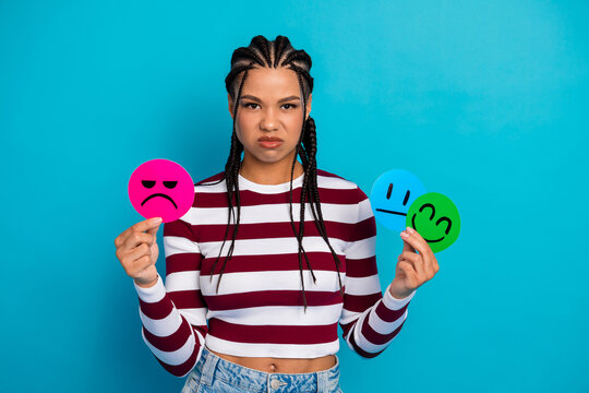 Young woman with braided hair holds sad pink face and blue and green smiles in bright studio wearing striped top against blue background