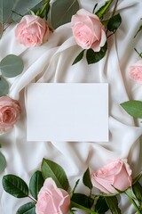 An elegant top view composition of pink roses and green leaves surrounding a blank white paper card on a draped silk fabric background for wedding invitations and romantic messages.