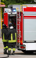 Two firefighters standing next to a red fire truck with a fire hose