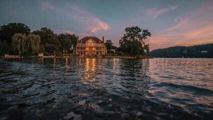 A lakeside house illuminated at sunset with trees and a calm water surface reflecting the sky and the building.
