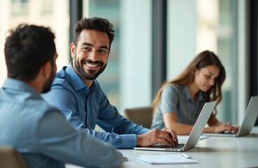Team works on laptops in open space office. Man smiles looks at camera. Coworkers use computers in meeting. Business people at corporate training in daytime workspace.