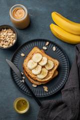 Peanut butter toast and banana on a black plate on a dark background with nuts